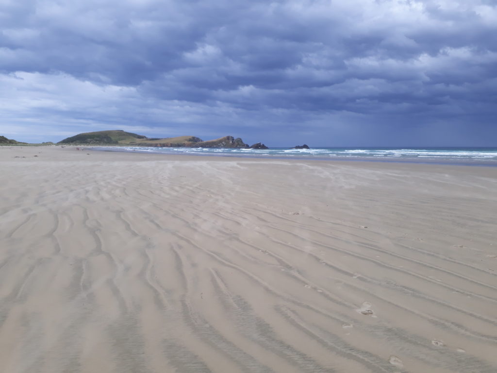 Cannibal Bay New Zealand, the beach in front and the ocean on low tide in background, dark sky with big blue cloudes
