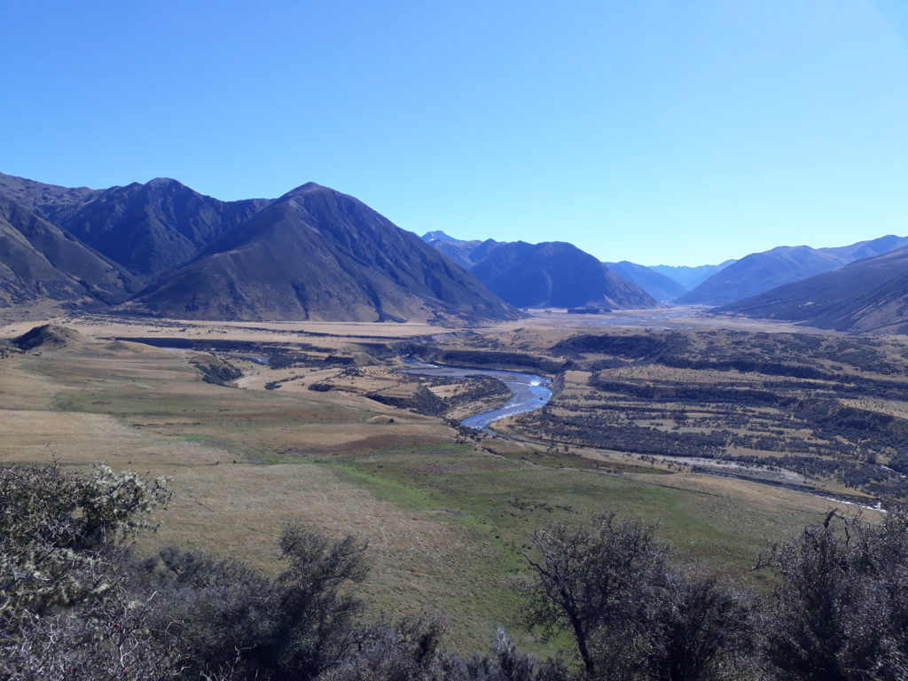 Blue sky, sunshine, view from a mountain into a valley, a river streams through the valley centered in the picture, left and right of the river tussock land and in the background grey and green mountains