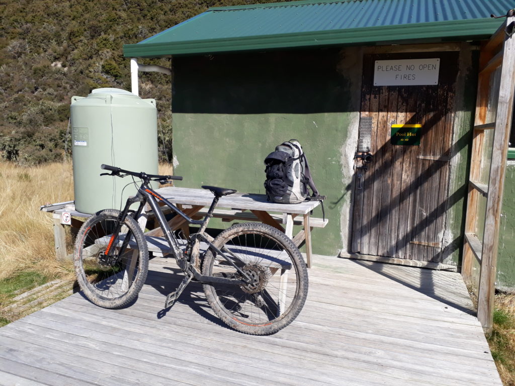 Blue sky, sun is shining, a green coloured hut along a tramping track, the hut covers two-third of the picture, in front of hut a small wooden porch with a wooden bench, a black mountainbike leaning against the bench and a small backpack standing on the bench