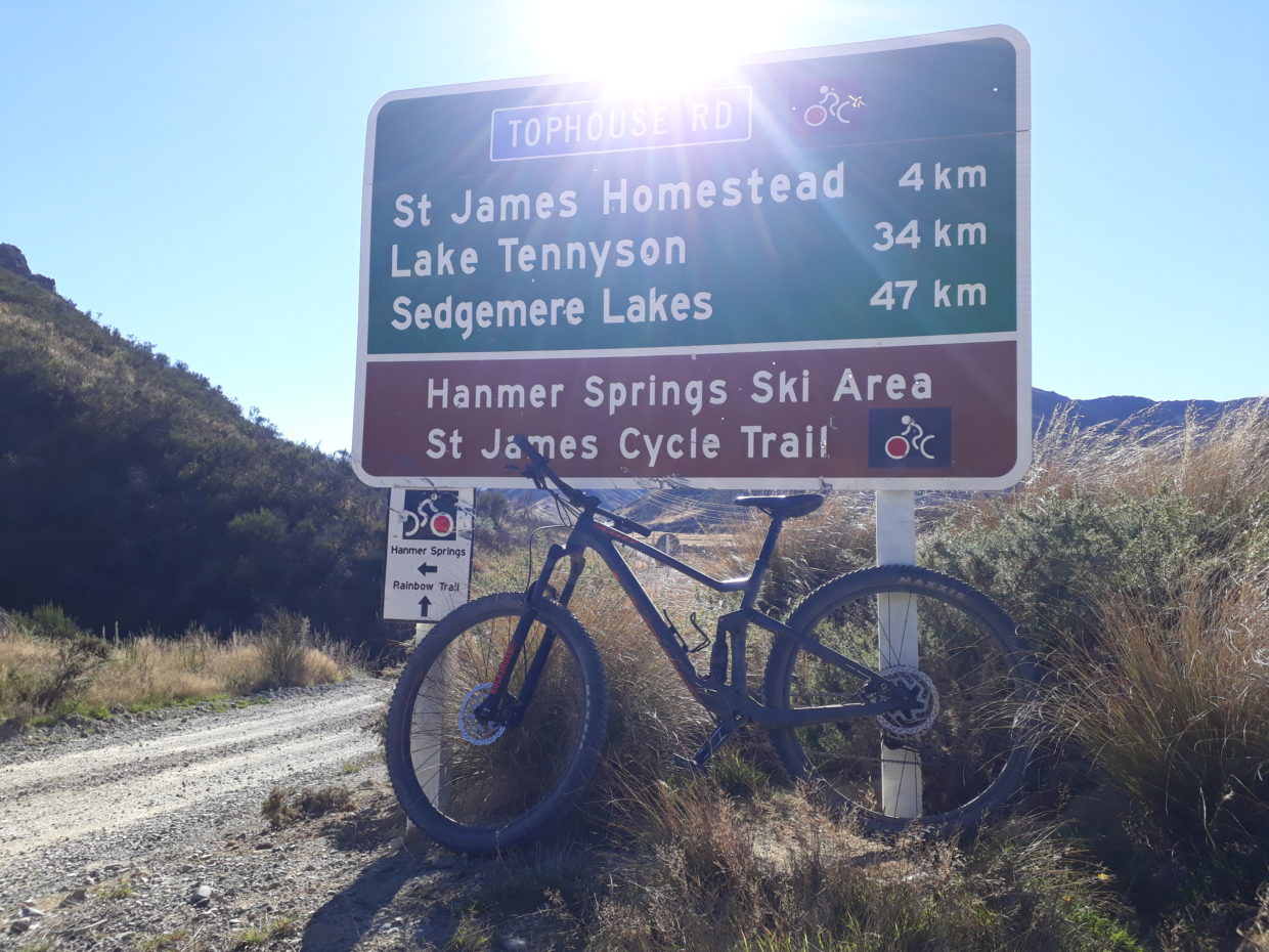 Blue sky, sun is shining, a big green signpost is centered in the picture giving distances to the next places and a hint about the Saint James Cycle Trail, the sun shines right above the sign so you can see some beams falling on the sign, a black mountainbike leaning against the signpost