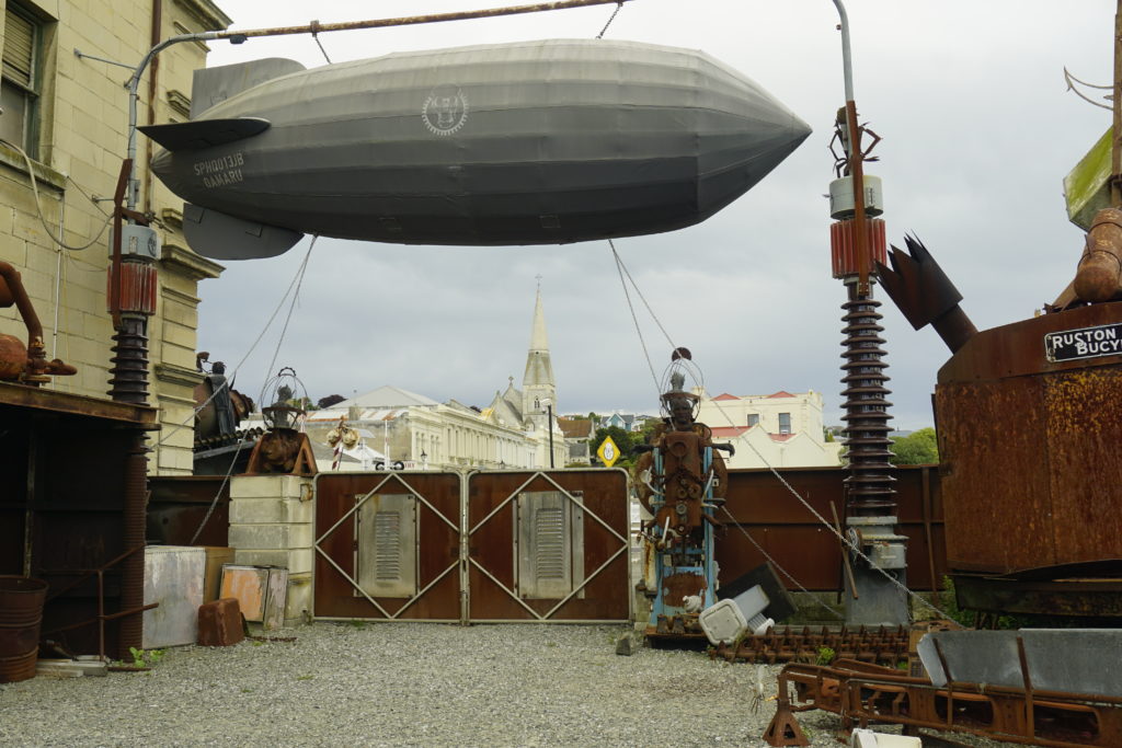 Steampunk Headquarter, scenery in backyard, left and right artificial metal made figure, in center a small sized zeppelin