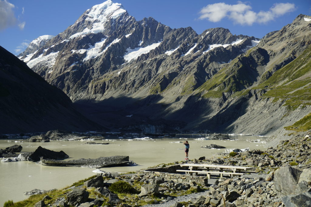Sunny day, blue sky, in front a lady standing on a bench, in background Mount Aoraki, a grey mountain with snow on top
