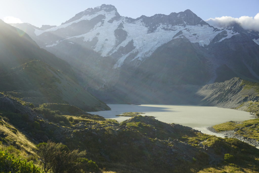 Sunny day, blue sky, sunbeams from left are shining in the picture, in front a grey glacier lake, background Mount Aoraki, a grey mountain with snow on top
