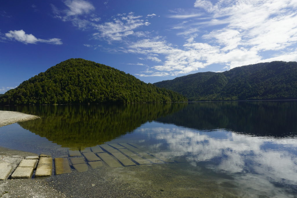 Blue sky, some white clouds, sunny weather, in the background some green bush covered hills mirroring in a lake which is in front