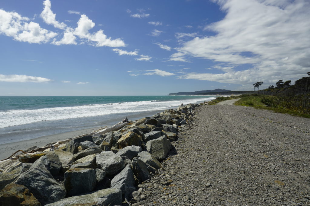 Blue sky with high clouds, sunny weather, the picture captures a coastal view with the ocean and beach on the left and a gravel road leading in the distance along the beach on the right, road and beach are separated by a rock wall