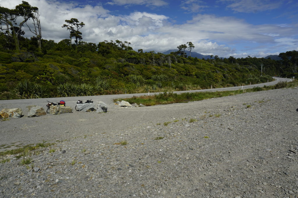 Blue sky with some white clouds, sunny weather, light winds, view on a coastal road from a beach, the road cut through a forest thus you have bushland next to the road, there is a beach access from the road secured by some big rocks, against this rocks are leaning two touring bicycles