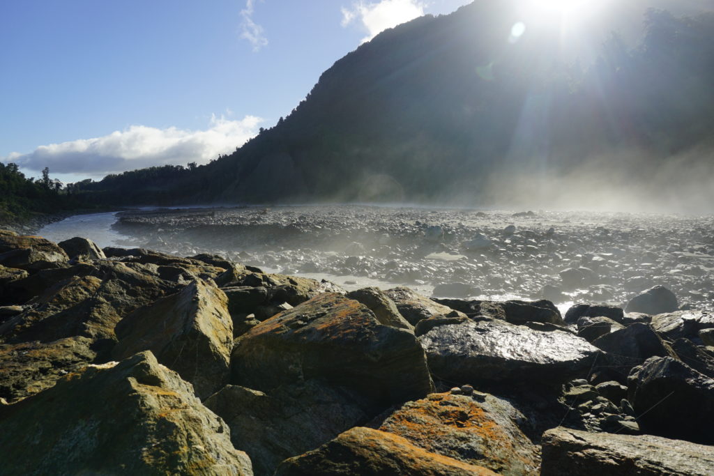 Blue sky, sun is shining, view on a river bed, the river is in the center of the picture, in the background is a mountain and some sun beams above shinig on the river, in front of the picture some big rocks from the river bed, haze is heated up by the sun and evaporates over the river bed