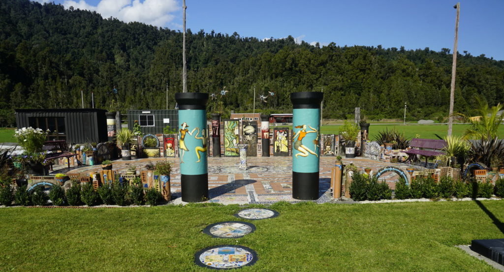 Blue sky, sunny weather, a picnic area in a village, a small court decorated with colorful tiles, the court is encircled by a small hedge, the area can be entered by a gate made of to big pillars with drawings on it showing people dancing, in the center of the court is a sundial