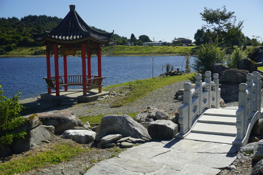 Blue sky, sunny weather, the pictures shows a picnic area on a small lake in a village, the area is decorated like a chinese garden, there is a small Chinese pavilion on the left and a tiny marble bridge with high and elaborated railings on the right