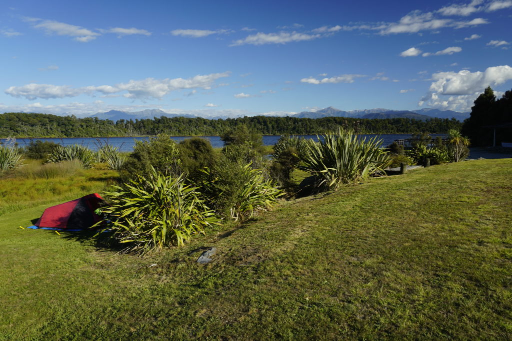 Blue sky, sunny weather, on the horizon mountains from left to right, in front a green lawn close to a lake with some single shrubs, next to a such a shrub is a black and red coloured tent, the lake in the background is blue and bounded by more bush in the background