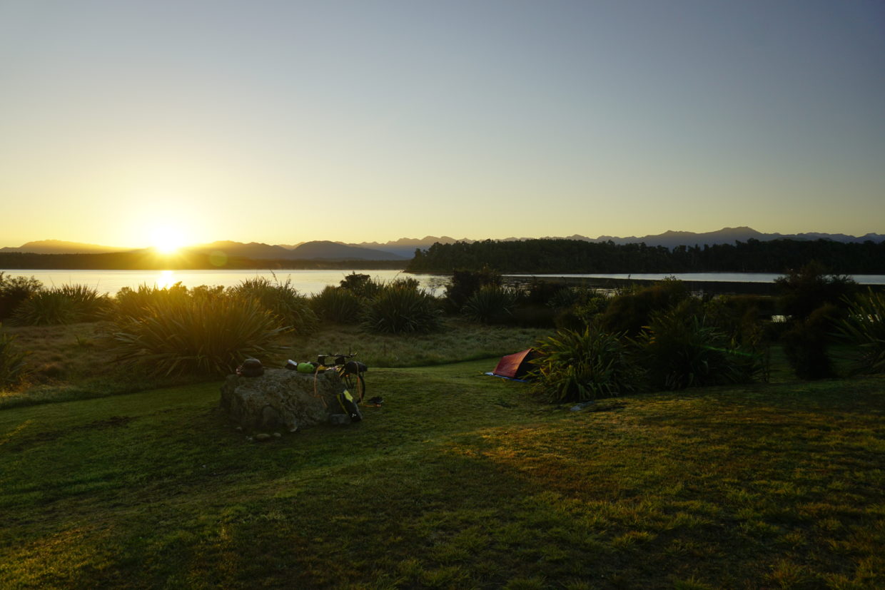A morning scenery, the sun arises just over the horizon, you can see first beams, the sky is clear, on the horizon mountains from left to right, in front a green lawn close to a lake with some single shrubs, next to a such a shrub is a black and red coloured tent, the lake in the background is blue and bounded by more bush in the background, next to the tent lies a big rock with touring bicycle leaning against it