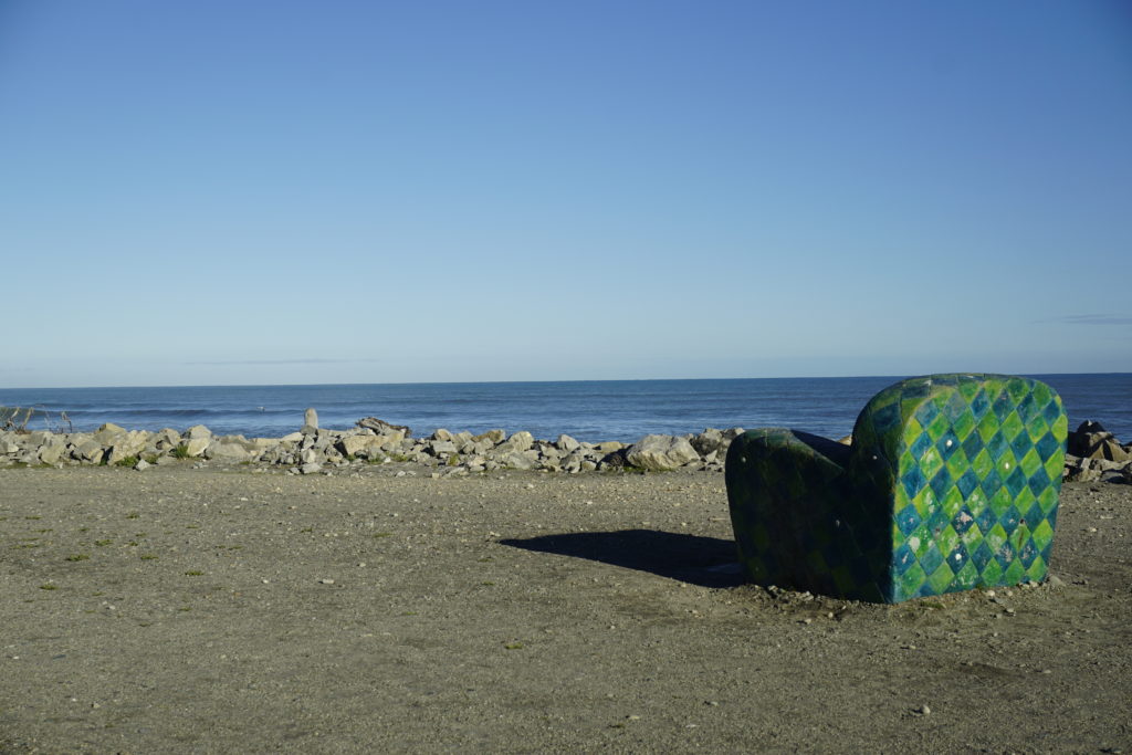 Blue sky, sunny weather, coastal view, the ocean in the distance, in front a gravel road with a big armchair made of stone, coloured with a green and blue tile pattern, the chair is directed to the ocean in 45° angle hence you can see its back and side, the sun shines from the back of the oberserver and throws a shadow in front of the armchair