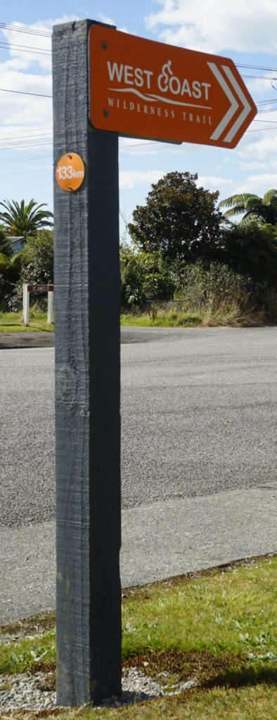 Blue sky, sunny weather, the picture shows a signpost in orange colour giving the direction of the West Coast Wilderness Trail in New Zealand