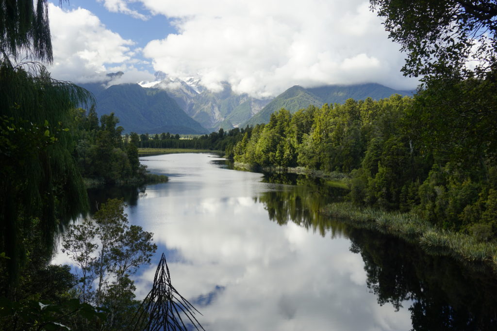 Blue sky, some white clouds, sunny weather, in the background a mountain skyline mirroring in a lake which is in front and encircled by bush witth tall trees