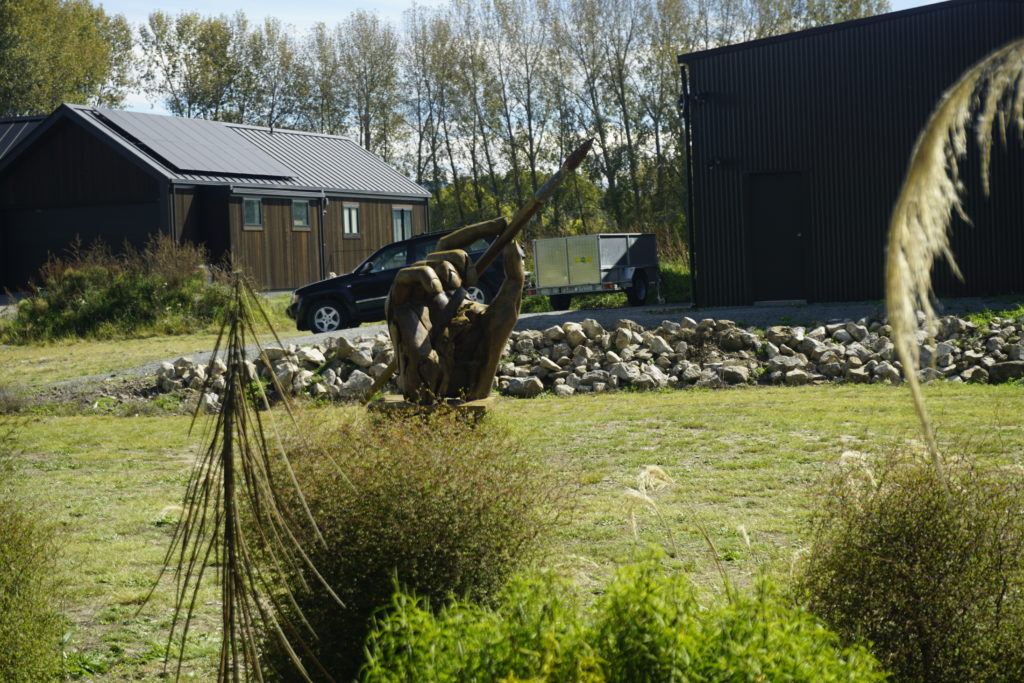 Blue sky, sunny weather, there is a sculptured hand standing on a pedestal and holding a paintbrush between thumb and index finger, the sculpture is located in a green garden