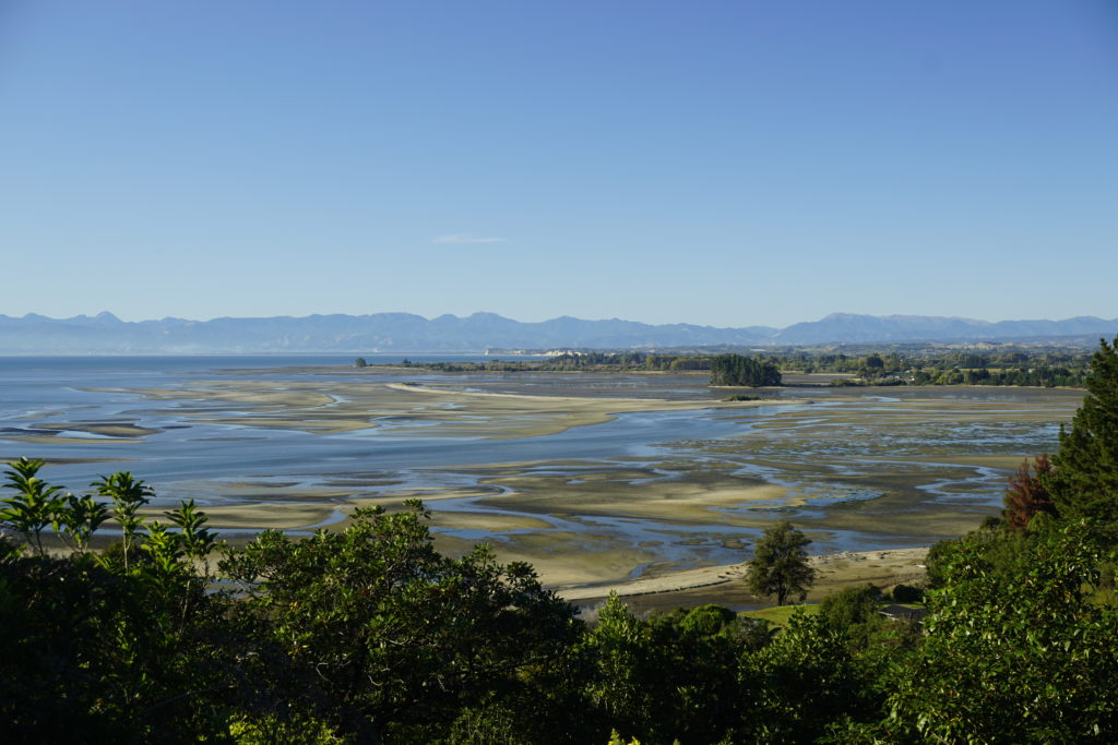Blue sky, sun is shiining, view from a hill, in front the slope of the hill covered with bush leading down to the coast, in the distance a mountain skyline with an water inlet at low tide in front, thus you can see water patches alternating with sandy patches