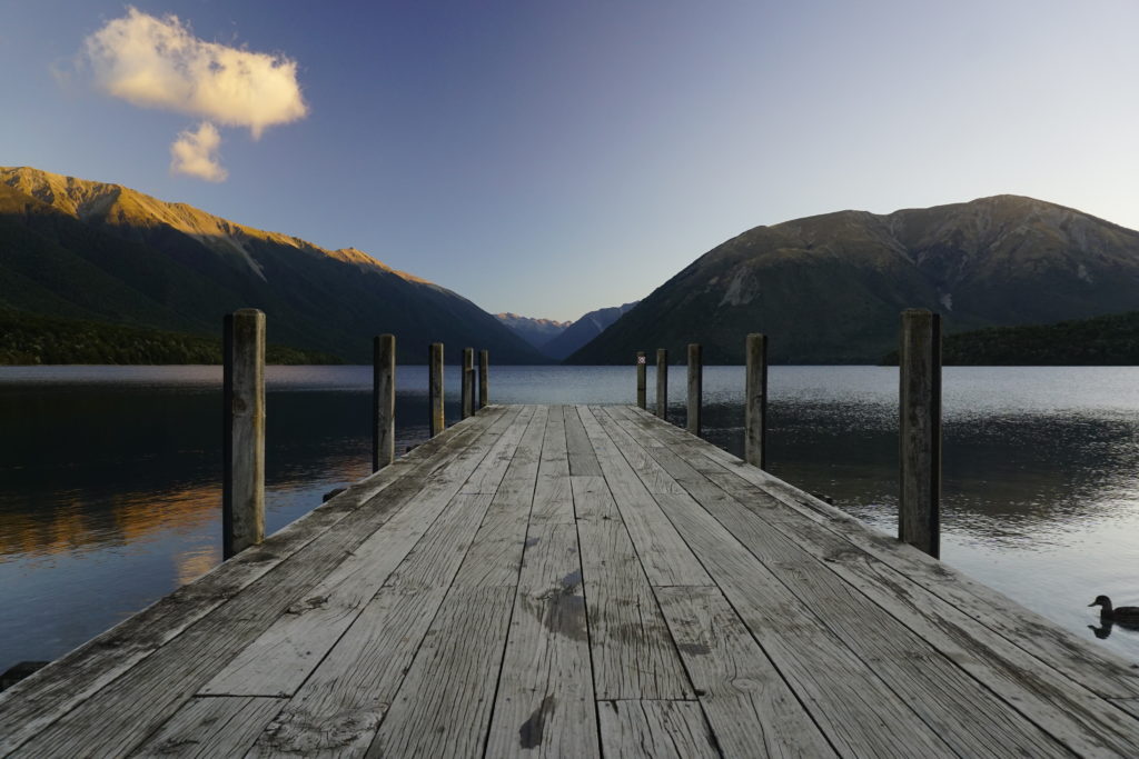 Sunny day on a late afternoon with sun slowly setting, blue sky, in center of the picture is a jetty leading into a lake, in the background mountains mirroring in the lake
