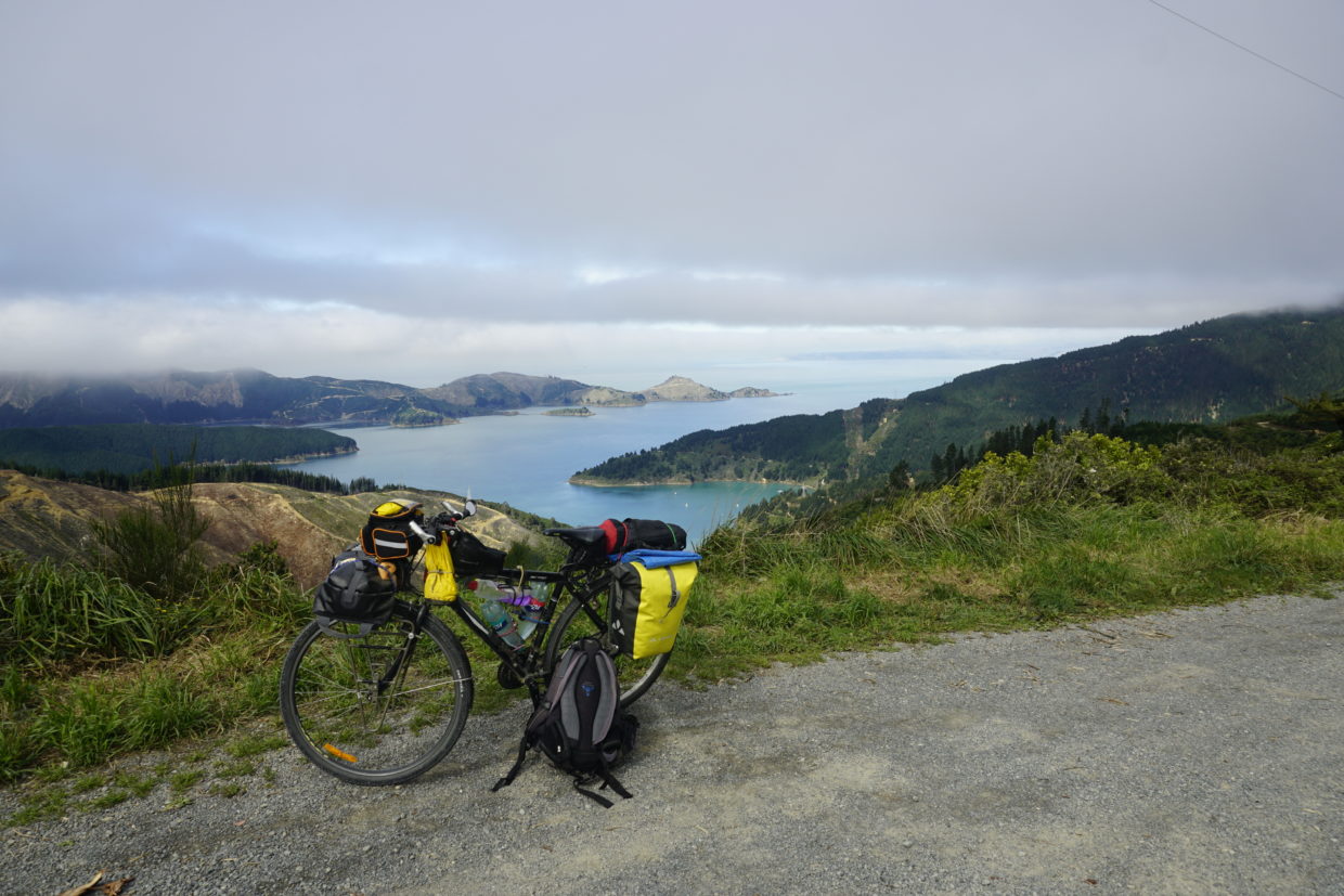 Blue sky, sun is shining, view from a hill, the picture shows an ocean inlet with the blue ocean in center touching the coast which kind of encircels the ocean, the coast is made of hills covered with forest and logging machines cutting off the trees, in front a touring bicycle
